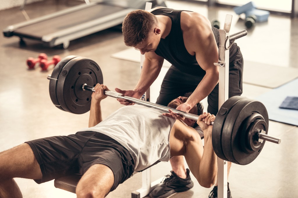 Athletic trainer assisting sportsman lifting heavy barbell in the gym