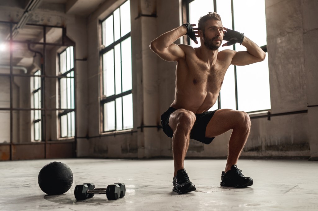 Shirtless muscular man with beard doing squats in a spacious gym