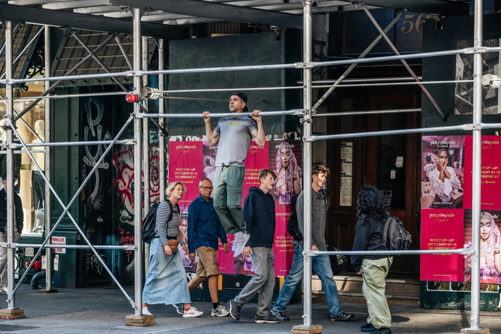 Mark Langowski does a pull-up on NYC scaffolding as people pass by on the sidewalk.