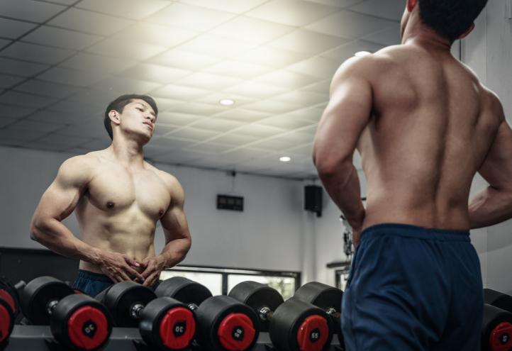 A man checks out his shirtless body in a gym mirror.