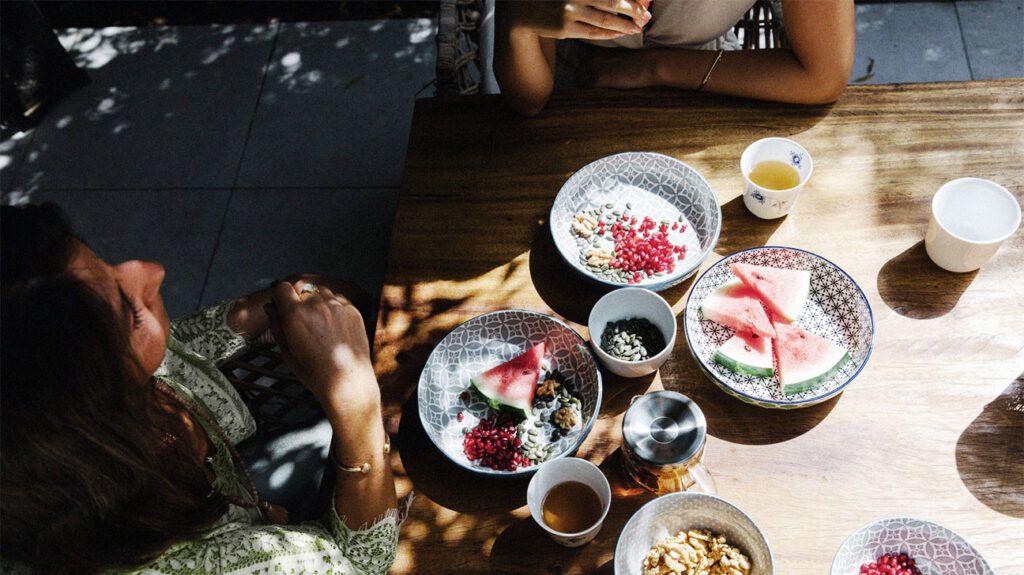 Two people having lunch outside with plates containing watermelon, pomegranates, and various seeds and nuts