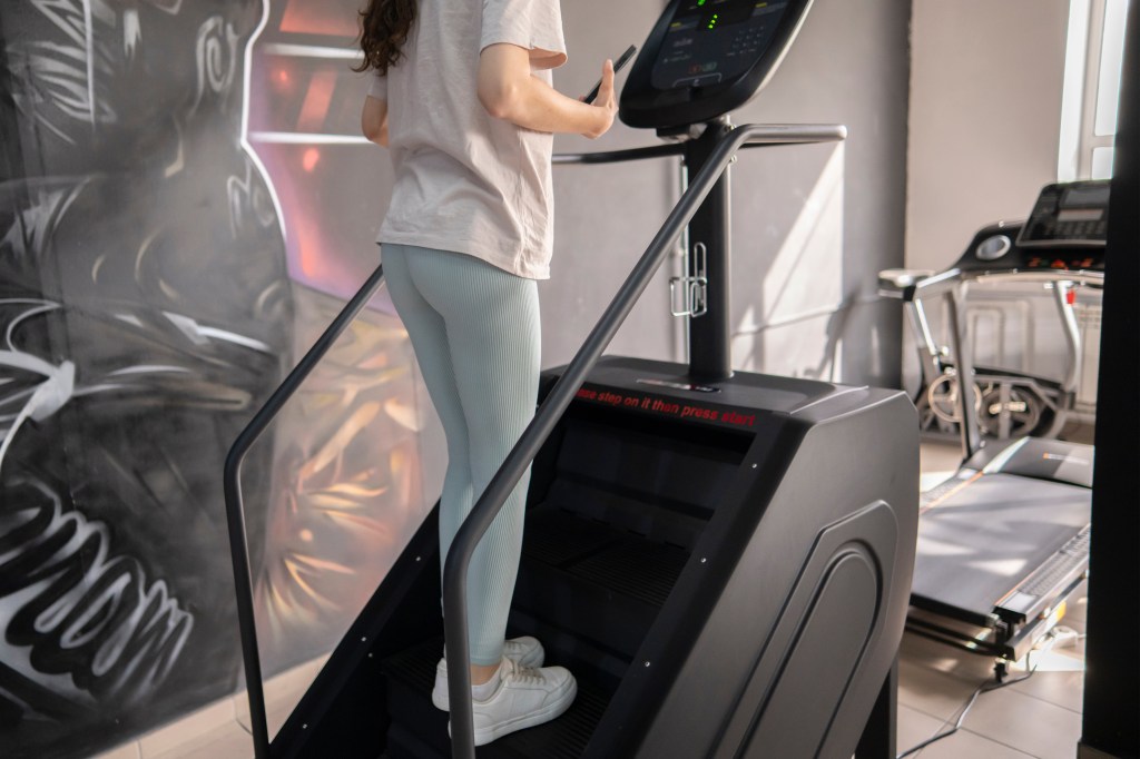 Young woman in athletic clothing exercising on a stair stepper machine in a well-lit gym with large windows and other equipment in the background