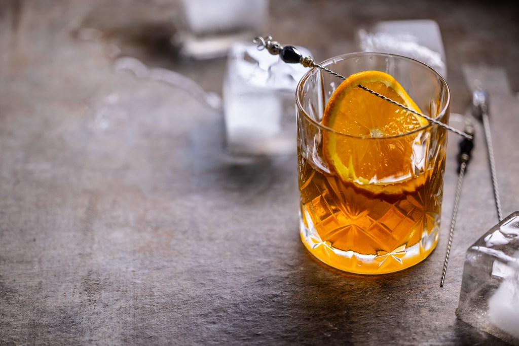 A glass of whiskey with ice cubes and a slice of orange on a stoneware table