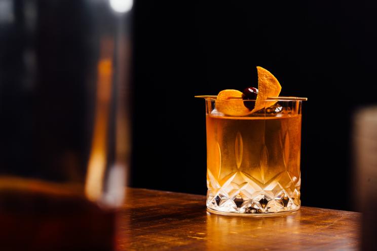 Old fashioned cocktail glass filled with orange liquid and garnished with orange chips, nestled between bottles in a bar