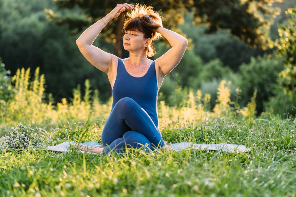 Middle-aged woman in blue sportswear performing yoga exercises on a grassy field during a sunset